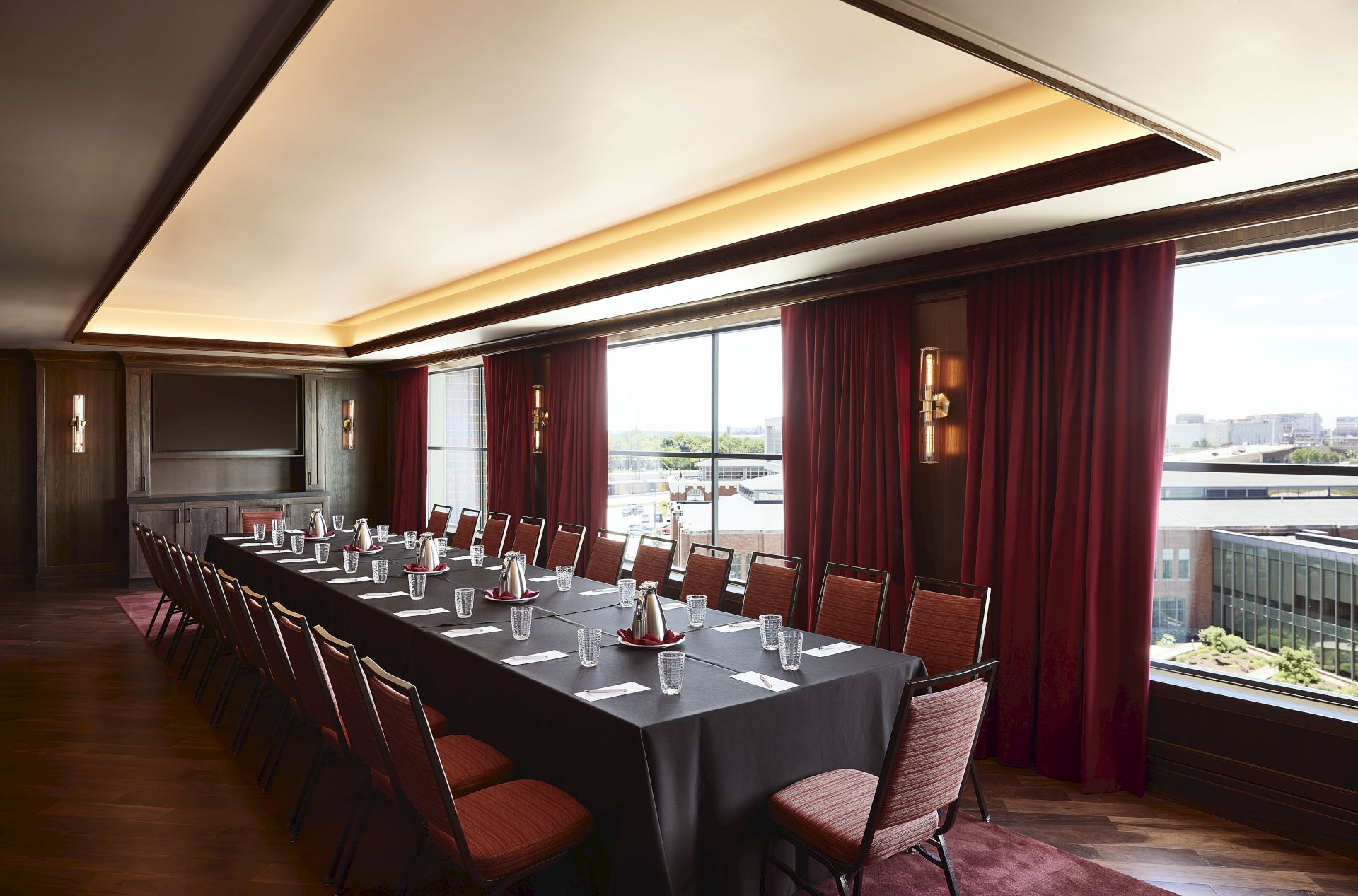 A bright conference room with a long table set for a meeting, red curtains, warm wall lamps, and large windows overlooking the city.