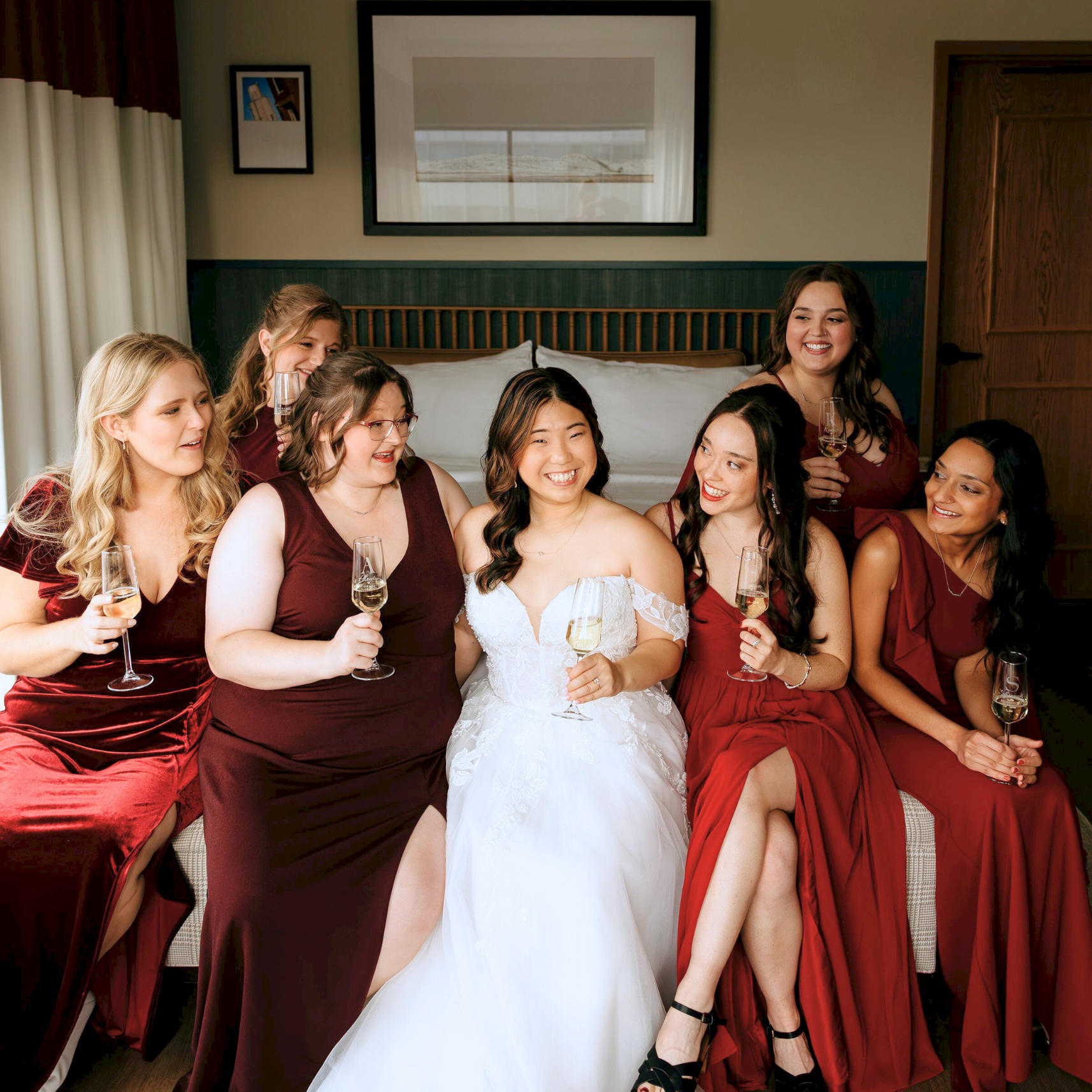 A bride in a white dress and her bridesmaids in burgundy gowns toast with champagne glasses in a cozy room.