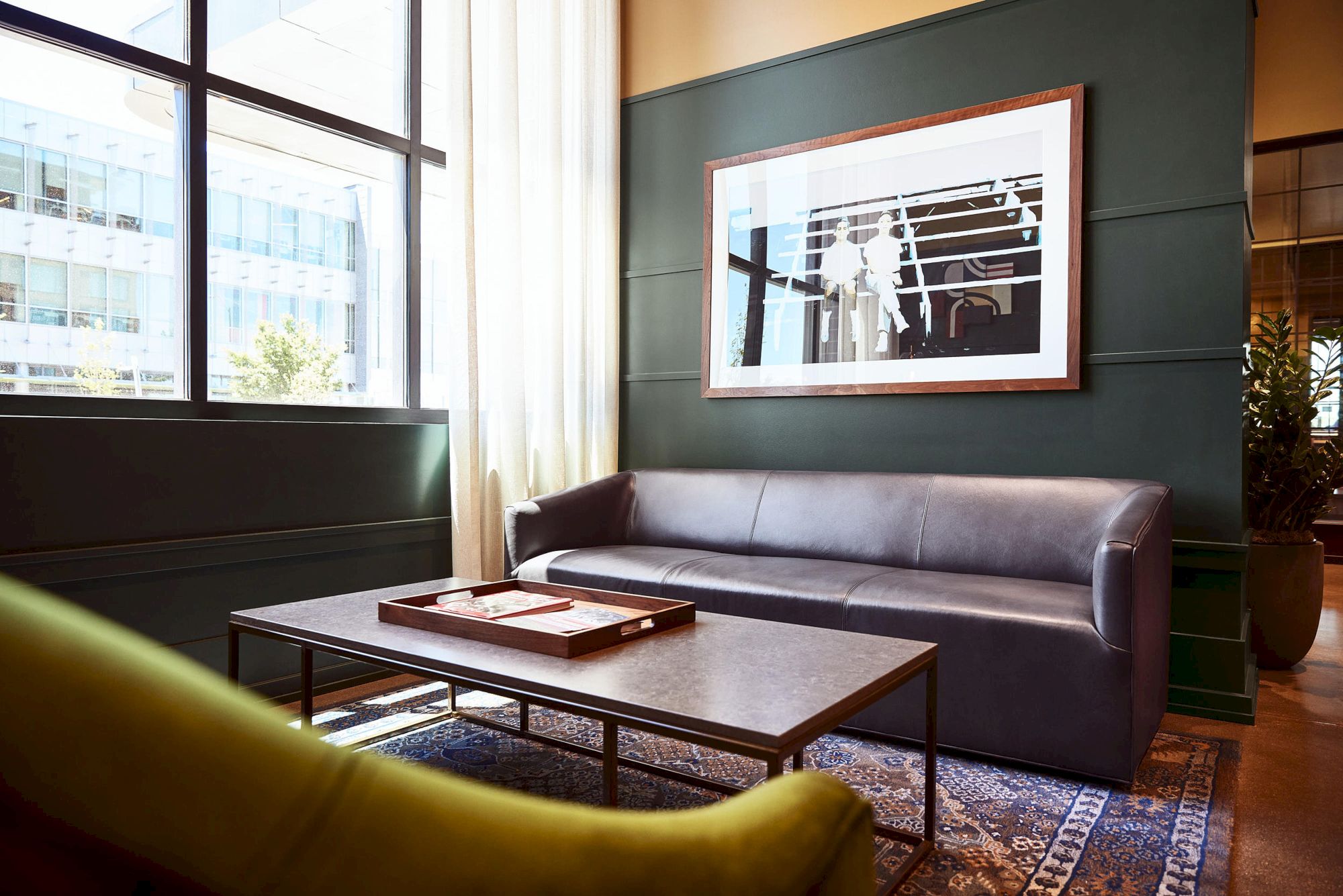 A modern lounge shows a leather sofa, coffee table, framed artwork, large windows, and a potted plant in warm light.