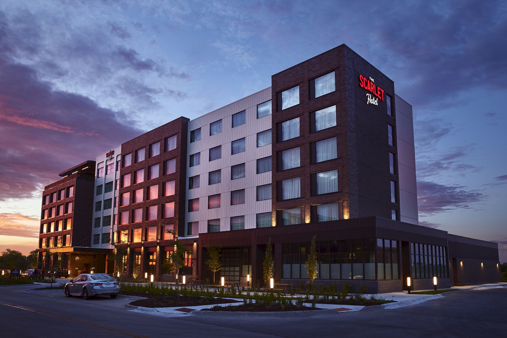 A modern building labeled The Scarlet Hotel at dusk, with glowing windows, trees, and a parked car on a street corner.