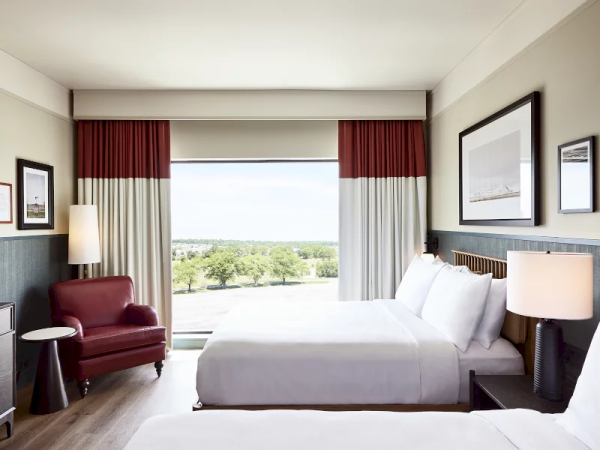 A bright hotel room with two beds, red-and-white curtains, lamps, framed photos, and a view of trees outside.