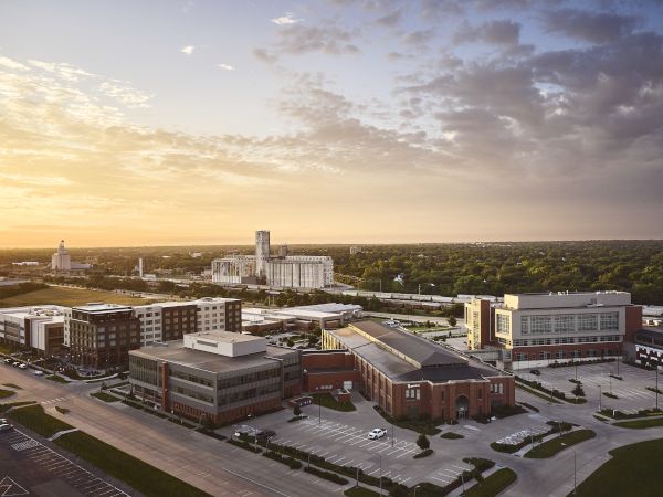 An aerial view of an industrial/campus complex with brick and concrete buildings, a silo tower, and a sunset sky with clouds.