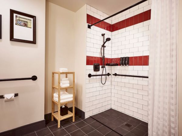 A modern tiled shower with a rain-style showerhead, dark floor, red-and-white accent tiles, and neatly folded towels in a wooden rack.
