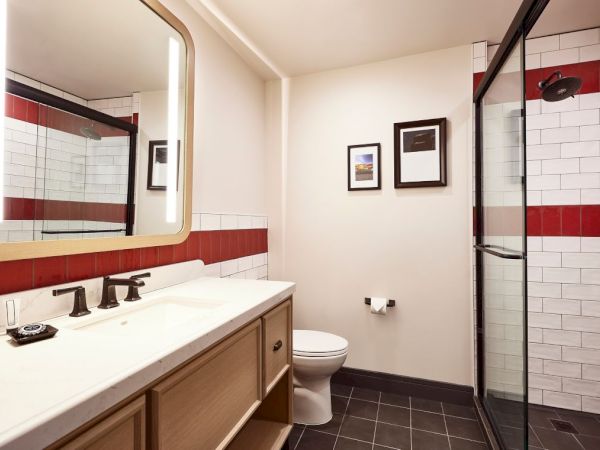 A modern bathroom with a white vanity, toilet, framed art, and a glass shower with tiled red-and-white walls.