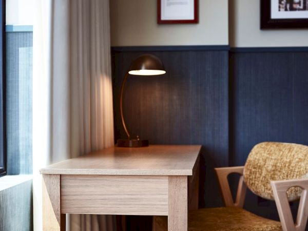 A cozy office nook with a wooden desk, lit lamp, and upholstered chairs by a window.