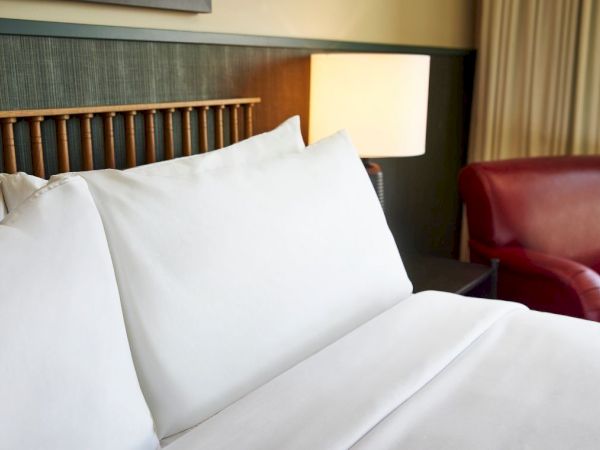 A hotel room scene with white bedding and pillows, a lit bedside lamp, and a red leather chair in the background.