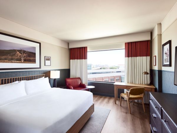 A modern hotel room with a large bed, red-and-cream curtains, seating area, window view, and a desk beside a dresser.
