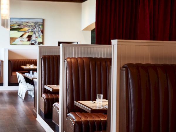 A cozy restaurant dining room with brown leather booths, wooden tables, and red curtains, viewed through an open doorway.