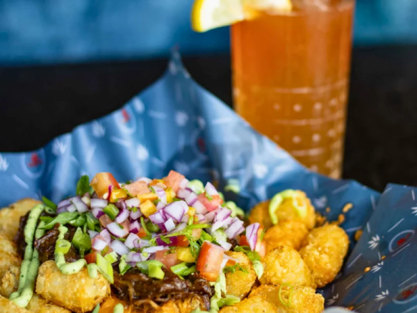 A tray of loaded fried chicken bites topped with salsa and chopped onions, with a glass of iced drink and lemon wedge.