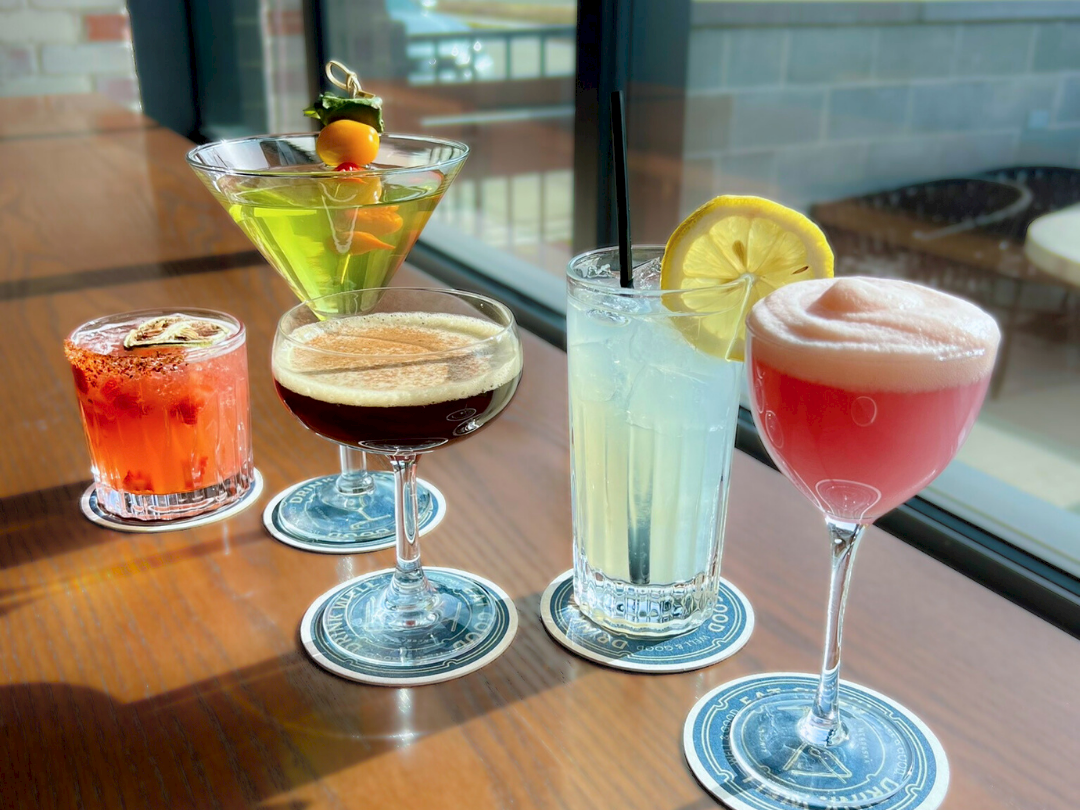 Four colorful cocktails on a wooden table&mdash;orange, green, dark red, and pink&mdash;garnished with citrus and served in stemmed glasses.