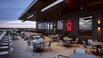 A modern rooftop patio with lounge furniture and wall art (a red devil-like logo) under evening lights.