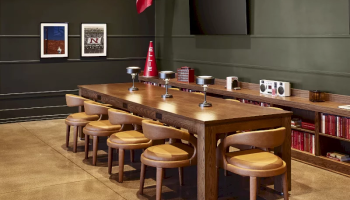 A meeting room with a long wooden table, cushioned chairs, wall shelves, framed photos, a red flag, and a TV on the wall.