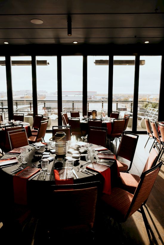 A set dining table with cutlery and glasses in a modern restaurant, viewed from inside toward large windows overlooking the city.