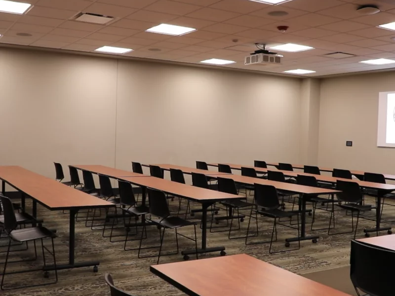 An empty conference room with rows of desks and black chairs, a projector screen, and ceiling lights.