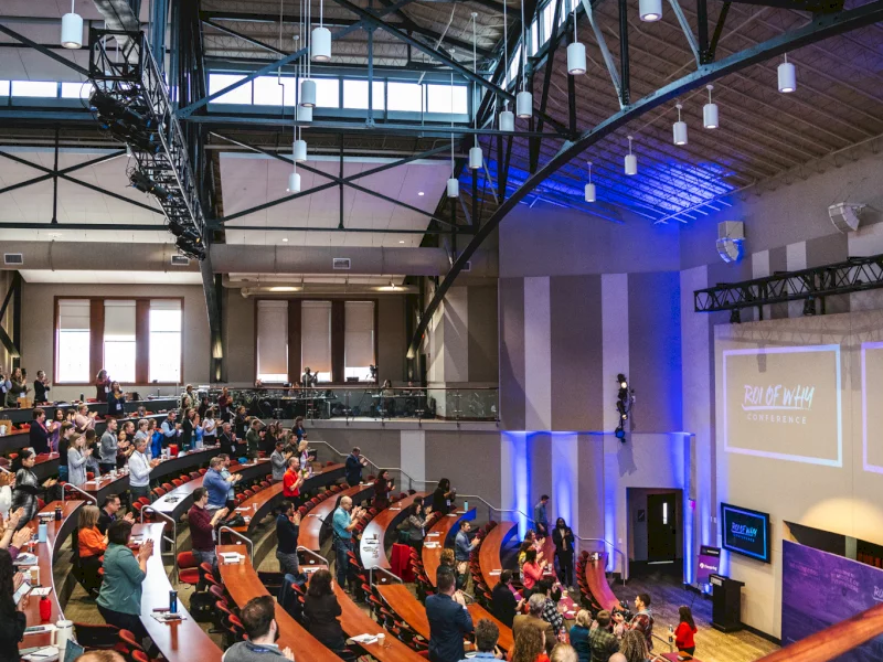 An indoor conference hall packed with attendees, speakers at a podium, and stage lighting shining over an event on a large screen.
