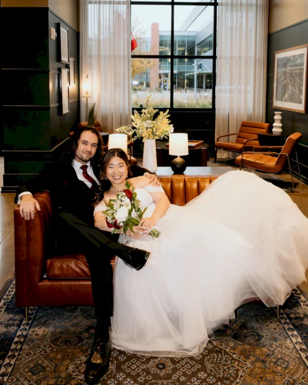 A wedding couple sits together indoors, the bride holding a bouquet and champagne as they smile in a warm lobby setting.