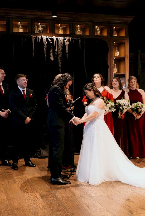 Bride in a white gown shares a moment at a wedding ceremony with groomsmen, bridesmaids, and guests in a lounge.