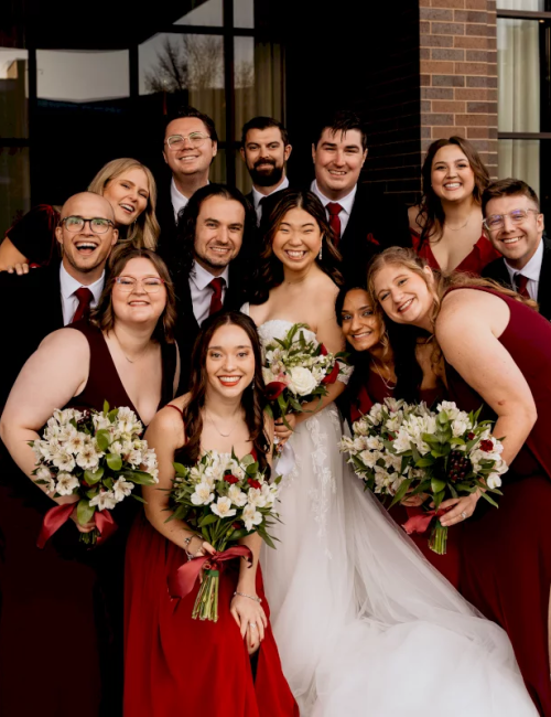 A wedding group photo with a bride in white and bridesmaids holding bouquets outside a brick building, smiling together.