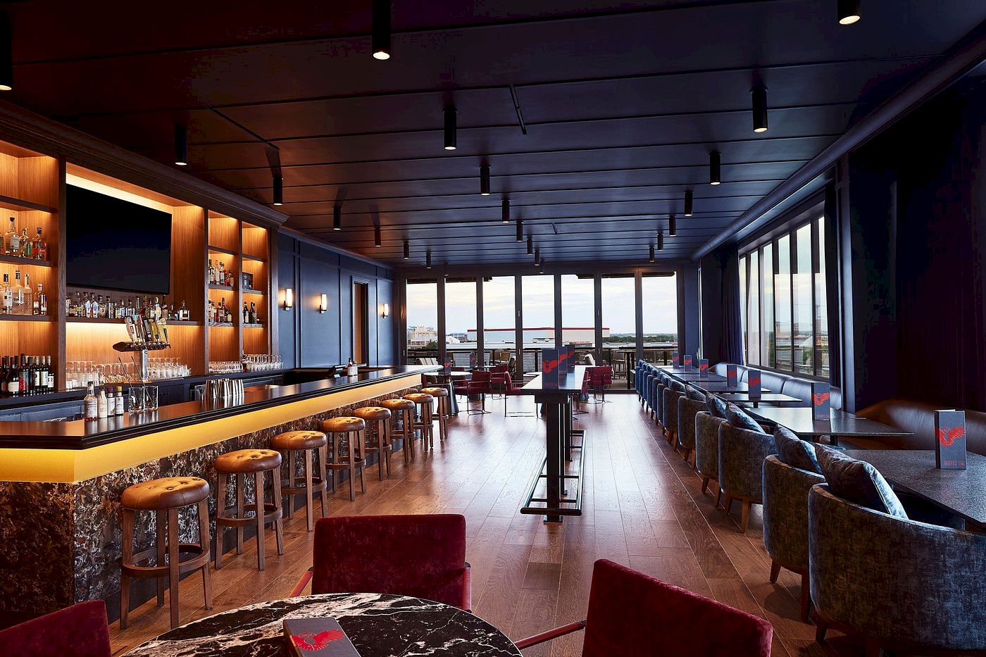 A sleek bar lounge with bottles, bar stools, plush seating, and floor-to-ceiling windows under pendant lights.