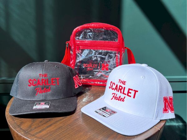 Two caps labeled &ldquo;The Scarlet Hotel&rdquo; (black and white) sit beside a red &ldquo;Scarlet Hotel&rdquo; bag.