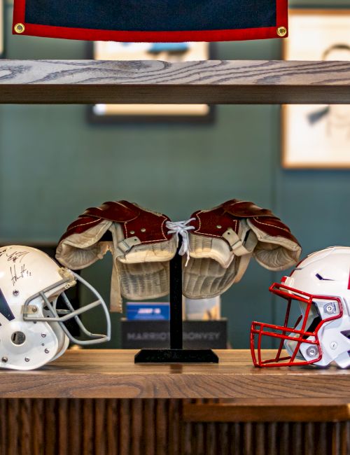 A display shelf with football helmets and jerseys, including a red-caged helmet marked with an &ldquo;N,&rdquo; plus a signed helmet.