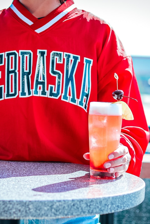 A person in a red Nebraska sweatshirt holds a clear iced cocktail with a cherry on a table.