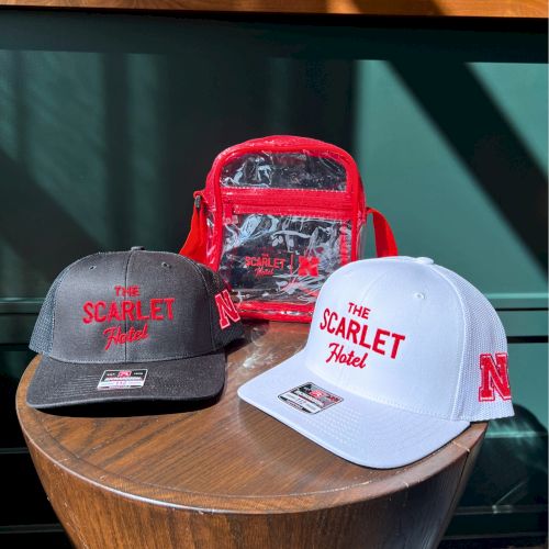 Two &ldquo;The Scarlet Hotel&rdquo; caps (black and white) sit beside a red &ldquo;Scarlet Hotel&rdquo; bag with the logo, on a wooden table.