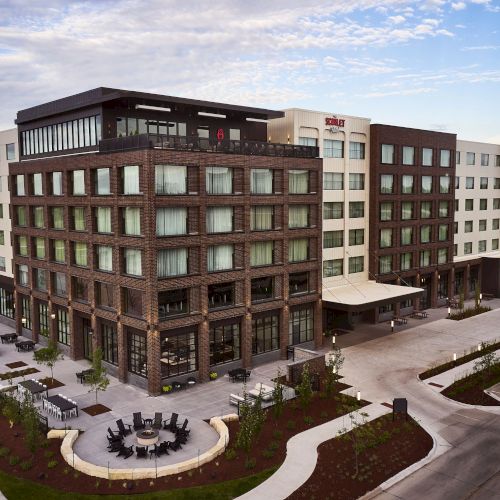 A modern multi-story brick hotel/residence building with a courtyard patio and cars under a blue sky.