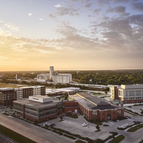 Aerial view of a university/industrial campus with brick buildings, parking lots, and silos at sunset under cloudy skies.
