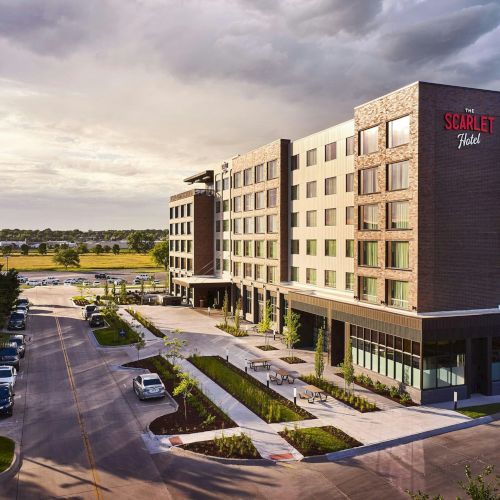 A modern hotel building labeled &ldquo;The Scarlet Hotel,&rdquo; with brick and glass walls, outdoor benches, cars, and a stormy sky.