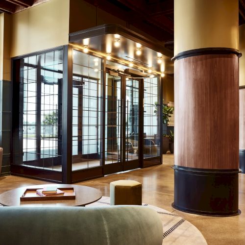 A modern office lounge with a couch and round table, floor-to-ceiling glass doors, warm lighting, and tall cylindrical wood columns.
