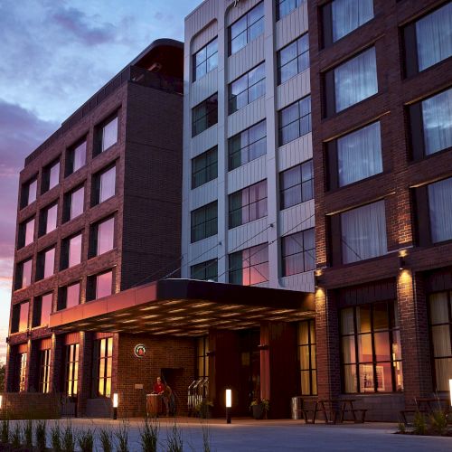A modern brick-and-glass hotel building with arched windows and warm lights at dusk, under a cloudy sunset sky.