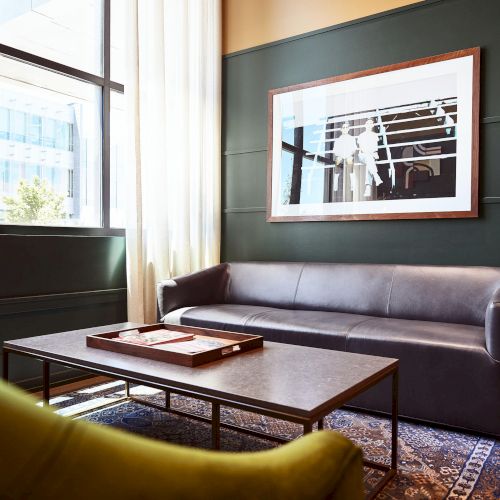 A modern lounge with a leather sofa, wooden coffee table, windowed view, and a framed artwork on the wall.