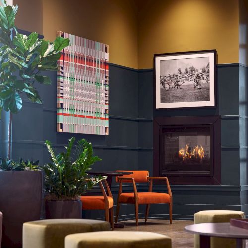 A cozy modern lounge with orange chairs, a lit fireplace, framed photos, and large potted plants by tall windows.