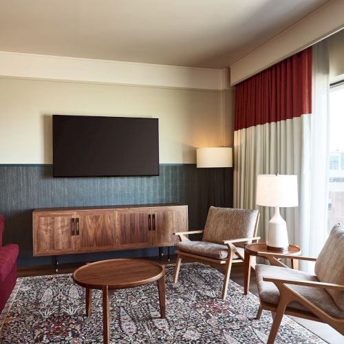 A modern living room with a mounted TV, red and white curtains, wood media console, lamps, and cozy seating by a large window.