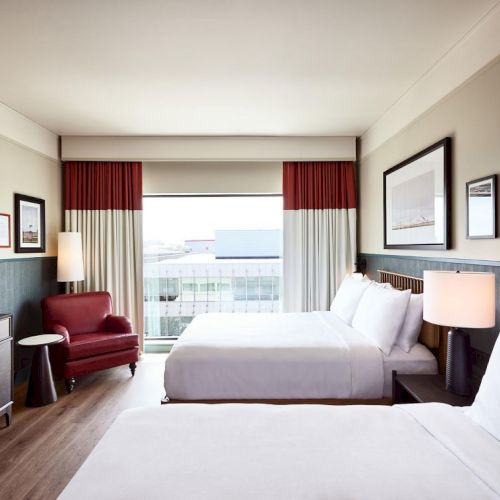 A modern hotel room with two beds, red curtains, lamps, and framed pictures, facing a window with city view.