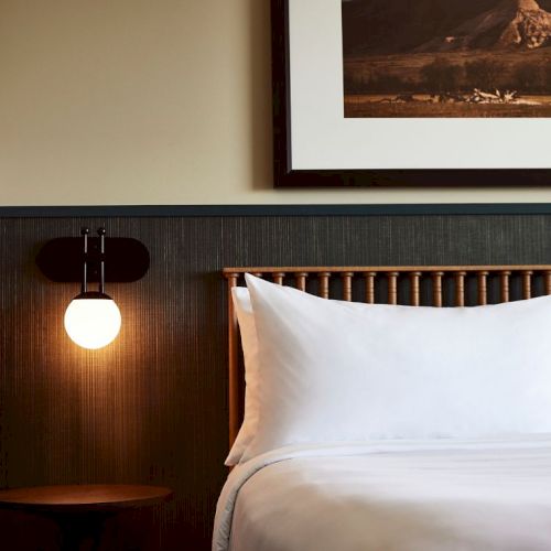 A cozy hotel room corner with a lit hanging lamp, a round side table, and a bed with white pillows.