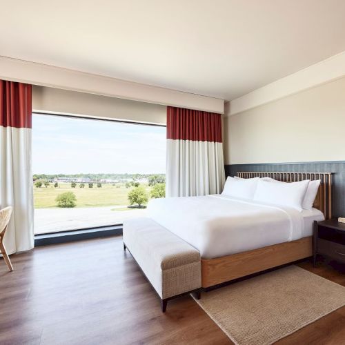A bright modern hotel room with a king-size bed, red-and-white curtains, a desk, and a scenic window view.
