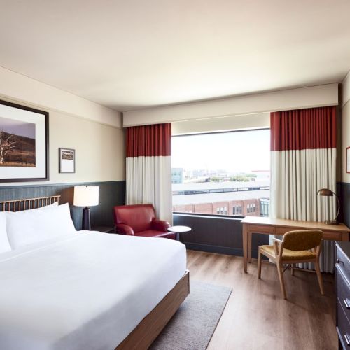 A modern hotel room with a large bed, red-and-white curtains, a seating area, desk, and framed wall art.