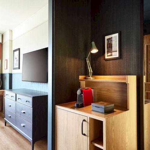 Modern hotel room with TV and dresser, plus a wooden desk niche holding a red espresso machine and lamp.