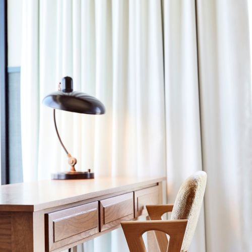 Cozy room: a wooden table with a lit lamp and a cushioned chair, framed by white curtains and red drapes.