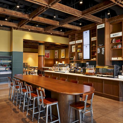 A modern caf&eacute; checkout area with a drinks fridge, coffee machines, shelves of treats/market items, and wooden bar seating.