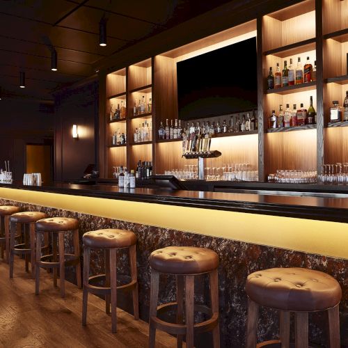A luxury bar interior with backlit shelves of liquor bottles, neatly lined glasses, and leather stools around a glowing counter.