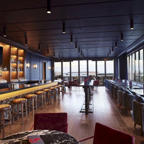 A modern hotel bar with a marble counter, stools, shelves of liquor, and lounge seating by large floor-to-ceiling windows.