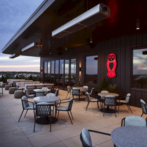 A modern rooftop patio with outdoor seating, retractable heaters, and a red owl sign at dusk.