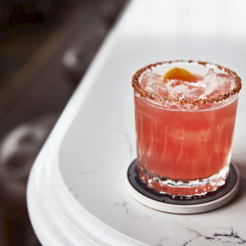 A frosty red-pink cocktail in a glass with icy garnish and a sugared/salted rim, served on a white plate.