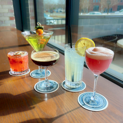 Four colorful cocktails with citrus garnishes in clear glasses on a table by a window, ready to sip.