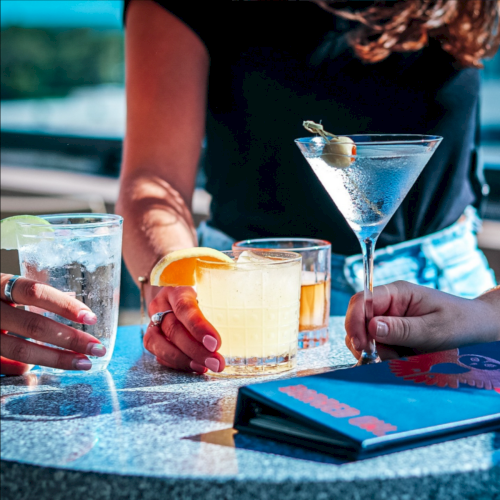 A group clinks cocktails outdoors: blue martinis and creamy drinks on a table with citrus garnishes and ice.