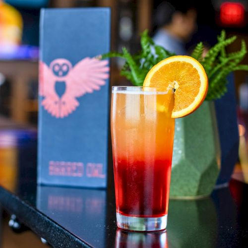 A tall glass with an orange-and-red layered drink, ice, and an orange slice garnish on a table.