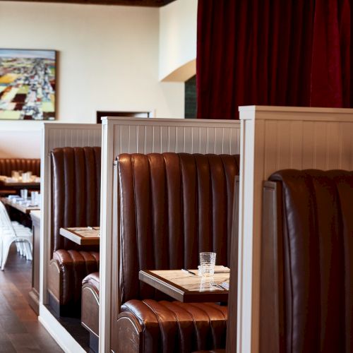 A cozy restaurant interior with brown leather booth seating, wooden tables, red curtains, and wall art on the back wall.